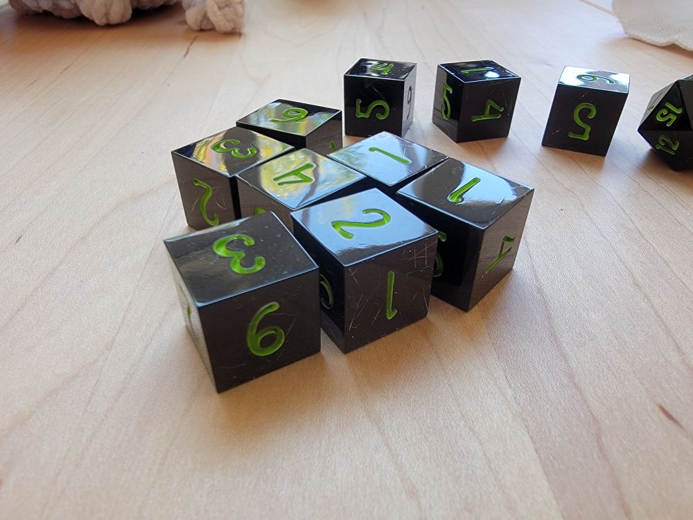 Ten six sided die on a light tan wooden table. The dice are almost entirely opaque black, just barely translucent enough to see metal threads winding through them. They are inked in a neon green color.