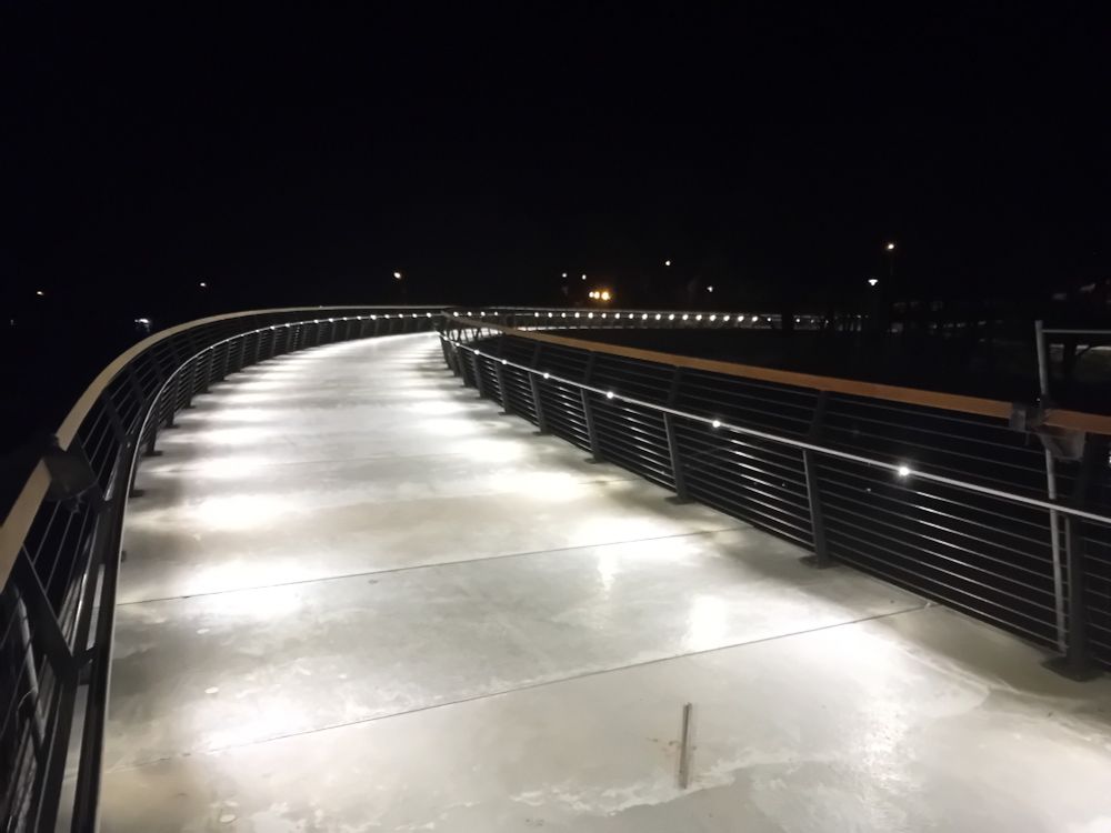 Gebogene Fußgänger- und Radfahrerbrücke mit LED-Beleuchtung im Handlauf.
