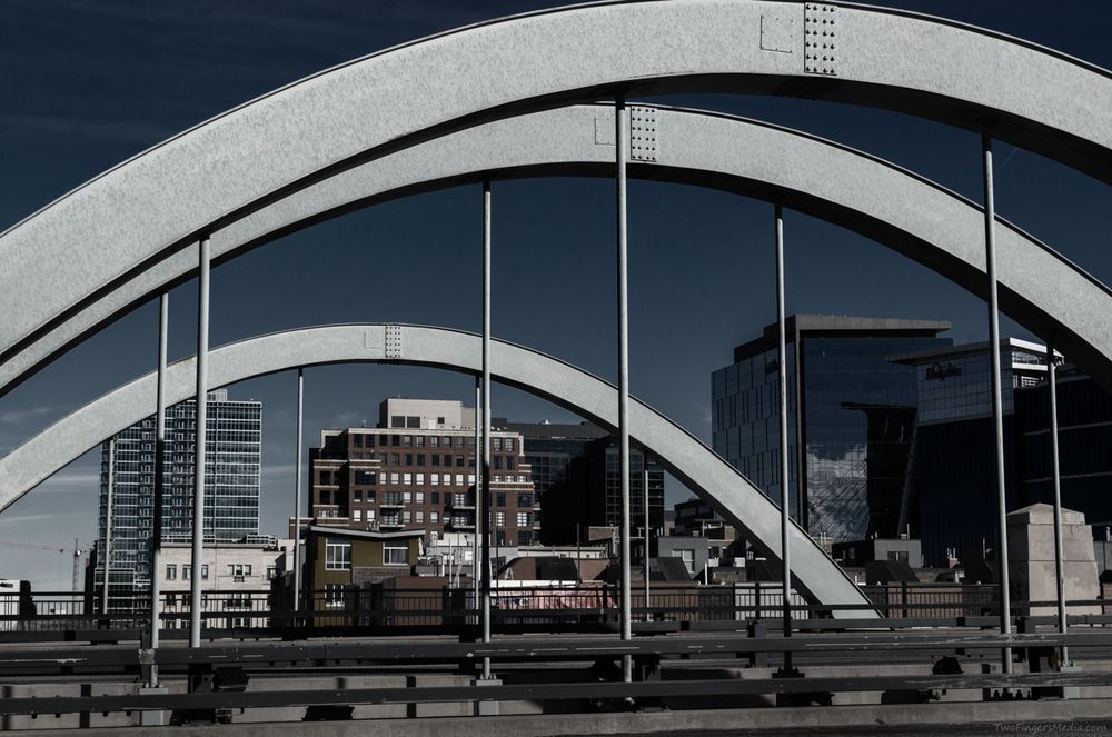 A photograph shows an urban scene framed by the curved steel arches of a bridge. The bridge's light-colored metal structure contrasts against a deep blue sky. In the background, a mix of glass buildings and older brick structures forms a diverse city skyline. The perspective emphasizes depth as the arches create a repeating pattern leading towards the cityscape.