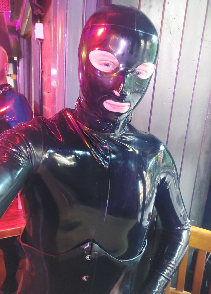 The rubber gimp is stood in a bar. It is wearing a rubber catsuit, hood, collar, and corset.
