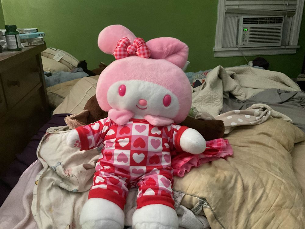 Image is a photograph of a large plushie of My Melody from Sanrio, the plushie is laying on a bed propped up by some pillows. She is a white rabbit with a pink hood, and is wearing red one-piece pajamas with hearts on them.