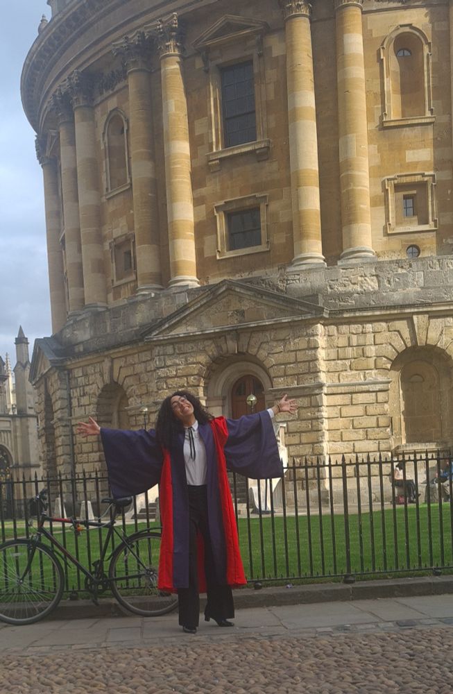 A young Black woman in red and blue academic gown celebrating inferring of an old yellow brown ornate building. 