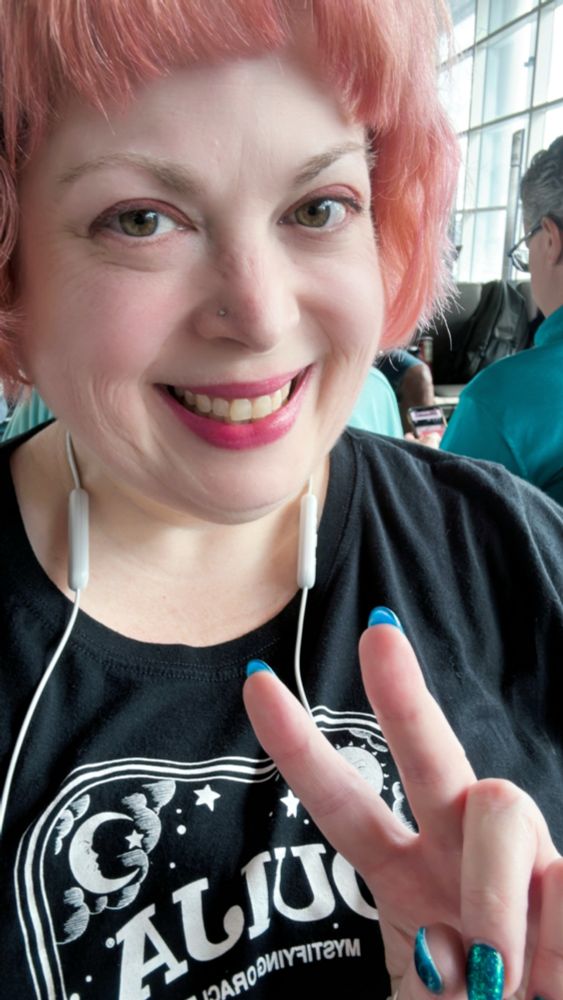 Pink haired lady making a peace sign and wearing a black Ouija t shirt. 