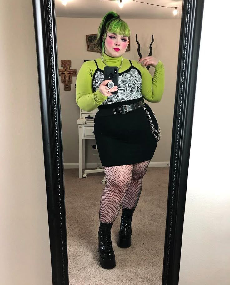 A cute chubby alt girl with green and black hair, wearing a green and black and white outfit, fishnets, and platform boots.  She's taking a picture with her phone in a mirror.