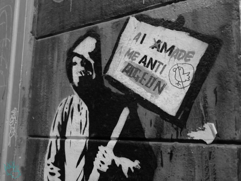 A B&W photo of graffiti on a wall - the picture is of a person wearing a hoodie and face mask, holding a sign that says "AI Made Me Anti Pigeon".  You can tell the sign part of the original graffiti has been defaced and the language has been changed.