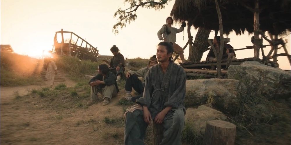 Scene from kdrama "The Murky Stream" set at dusk, showing characters sitting on rocks and tree stumps, dressed in rags. The surrounding area is dusty and there is  a shelter-type structure in the background.