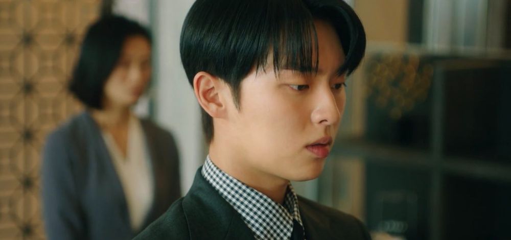 Close up of main male character Ban Juyeon, in Kdrama "My Dearest Nemesis" wearing a dark grey suit with the collar of a checked shirt visible. He has short black hair, parted in the middle.