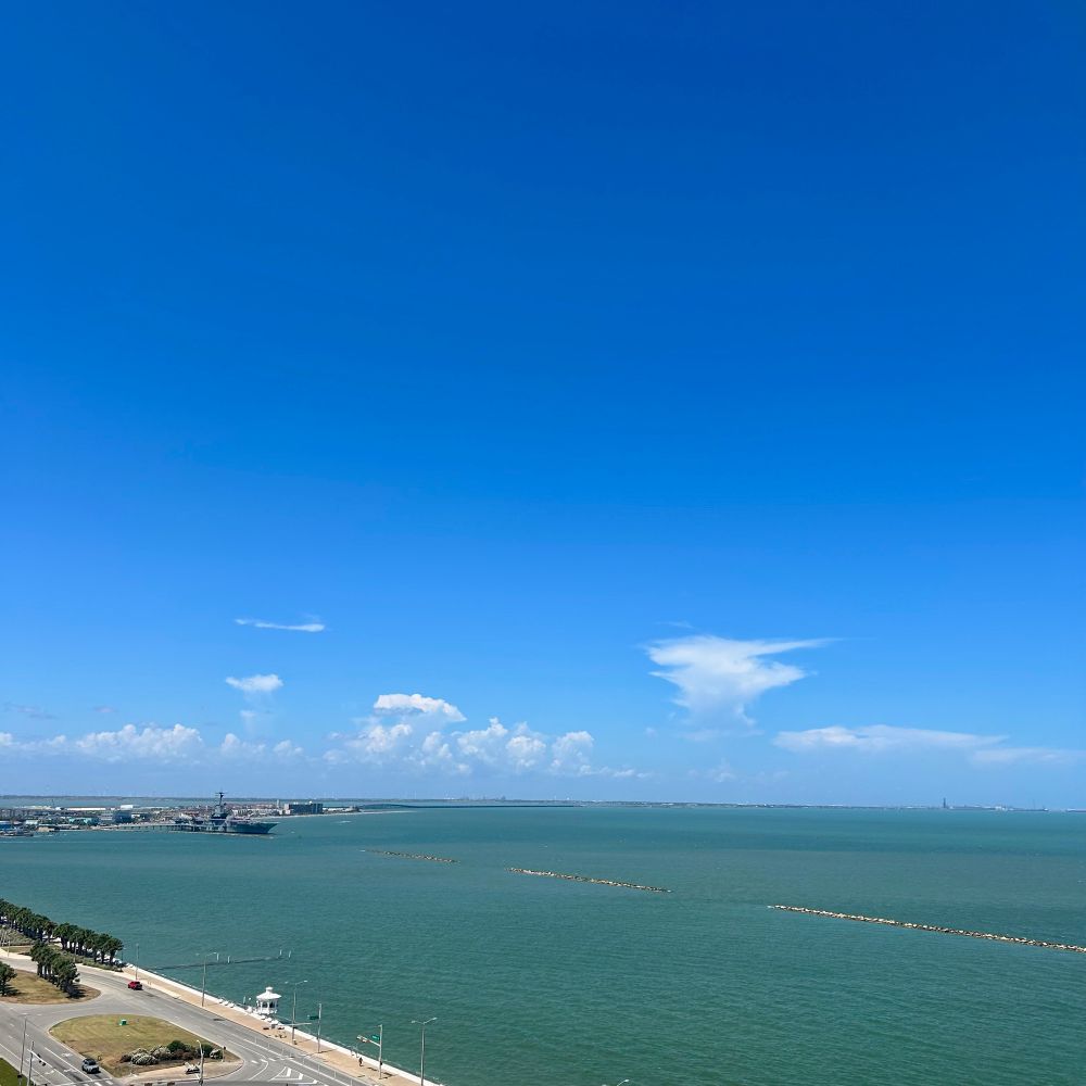 photo of the view from the balcony of my hotel room in corpus christi. the water of the gulf of mexico is visible and is a slightly brownish blue green hue. there is a large expanse of blue sky with fluffy clouds near the water fading into a pure blue sky above that takes up about half the frame. 