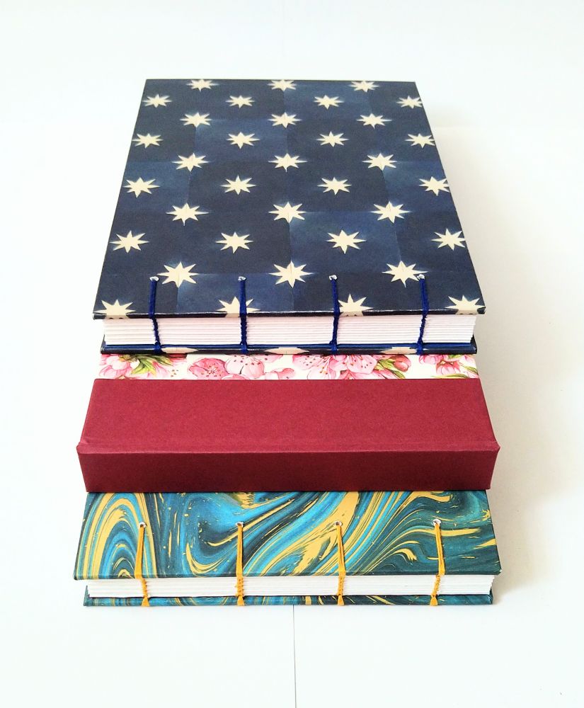 A photograph of three landscape format sketchbooks against a white background, spines towards the camera, two with open Coptic-sewn spines, one with a hard spine. One is blue with white stars, one is pink and floral with a red spine, and one with a blue and gold marbled cover.