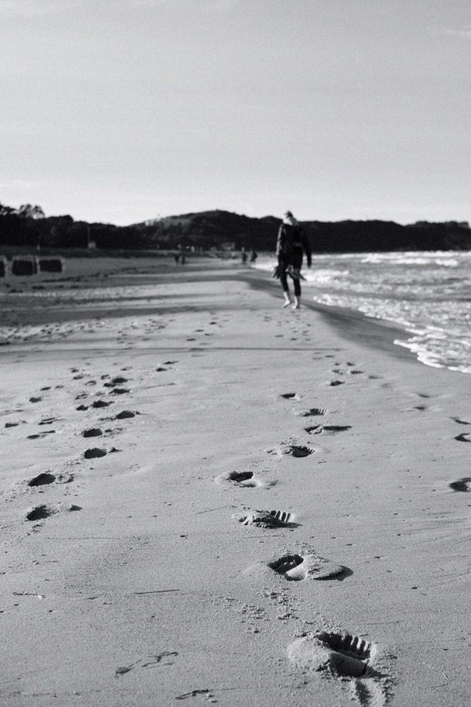 Fußspuren im Sand, am Strand. In ca 20m Entfernung vom der Aufnahme, eine Person die die Fußstapfen erzeugt. Das ganze in schwarz weiß gehalten. 
