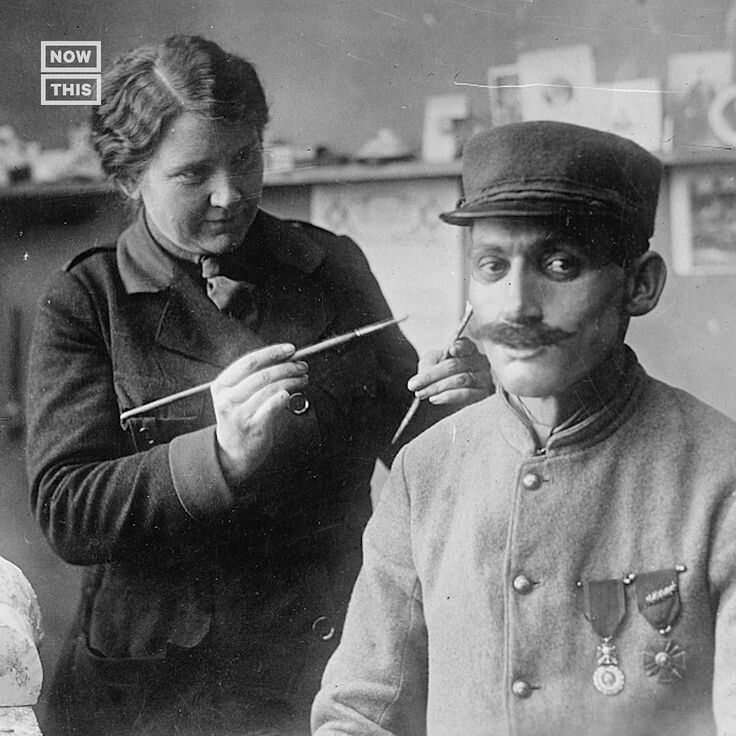 A black and white photo from the First World War. There is a soldier seated in the foreground. He is wearing a metal mask that has a mustache. Behind him is a woman holding a paint brush. This is Anna Coleman Ladd, the artist who created the mask.
