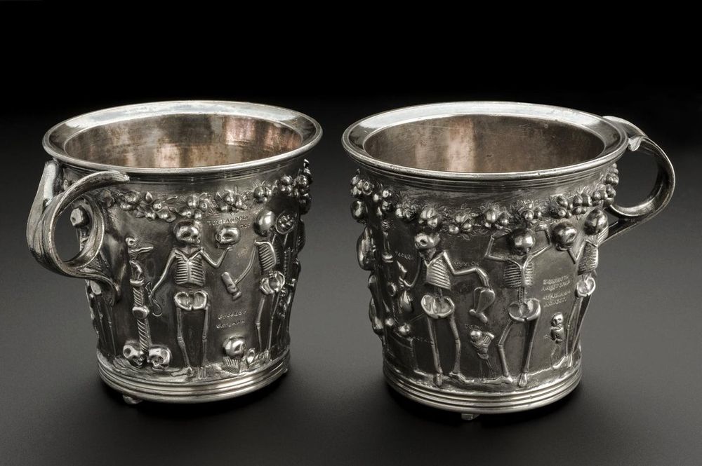 Two small silver cups with ornate handles. There are decorative skeletons all around it. 