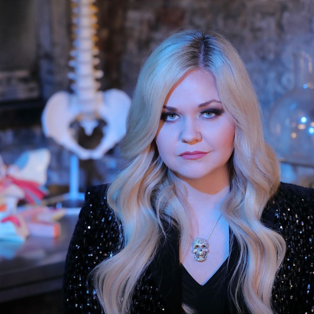A photo of Lindsey with long blonde hair wearing a skull necklace. The background is vaguely medical. 