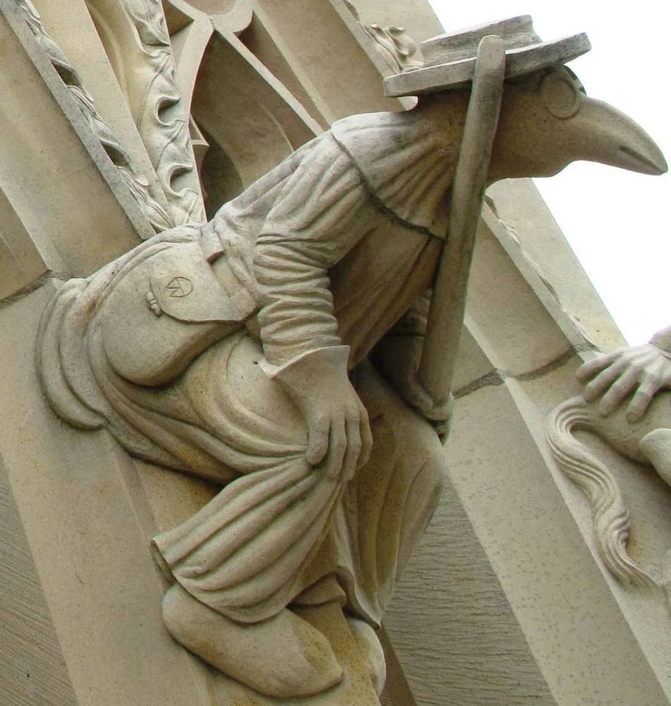 A carving of a plague doctor wearing a beaked mask, long robe, gloves, and a brimmed hat. He's also holding a pole, and he has a little satchel. 