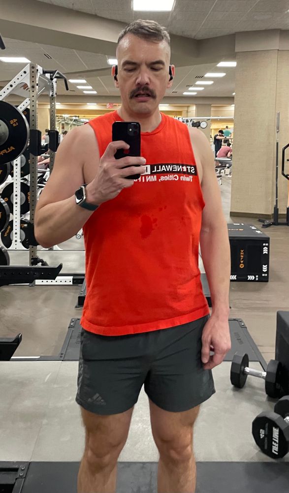 A man taking a selfie in a mirror at a gym. He is wearing a red cutoff t-shirt and gray shorts.