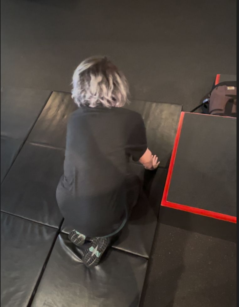 A woman with hair that is 2/3 bleached white, 1/3 grown out dark brown kneeling on a padded floor. Still learning strategies for getting to my feet.