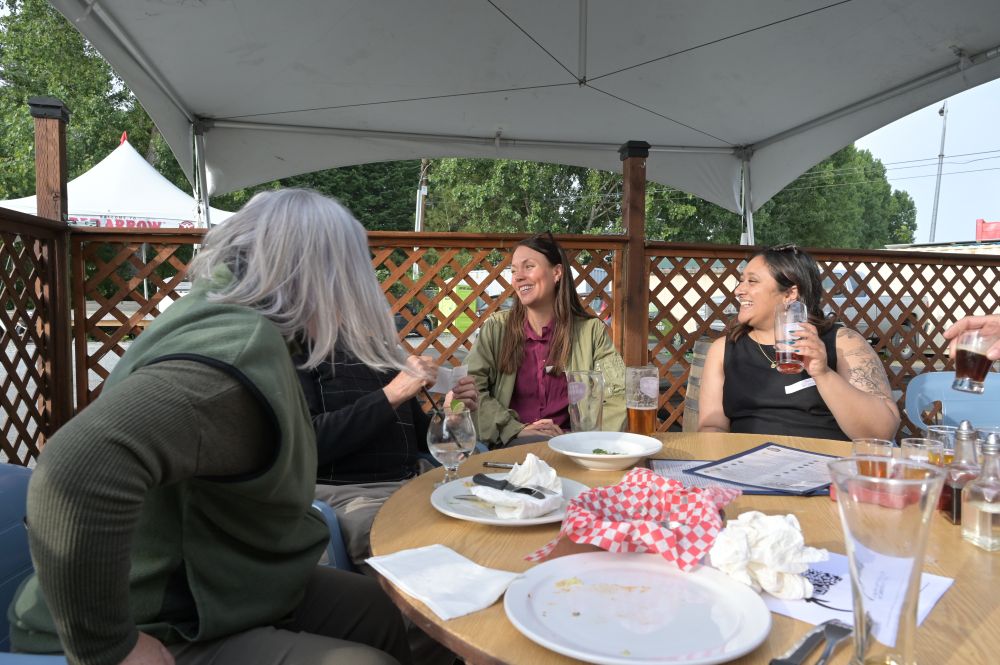 A group of people sit aroud a patio table holding beer laughing, with plates of food on the table.