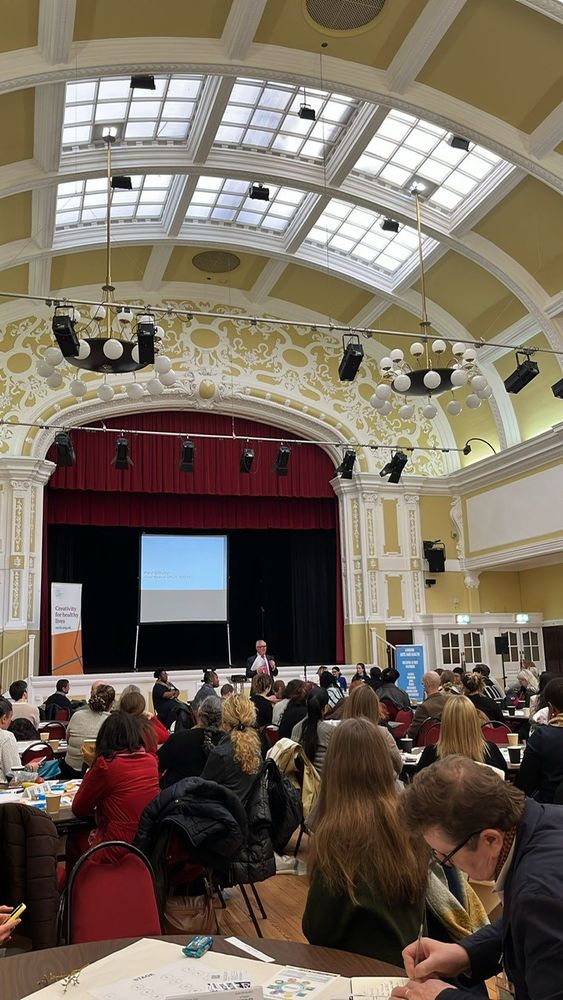 People sitting in a large hall with a man speaking in front of a stage. 