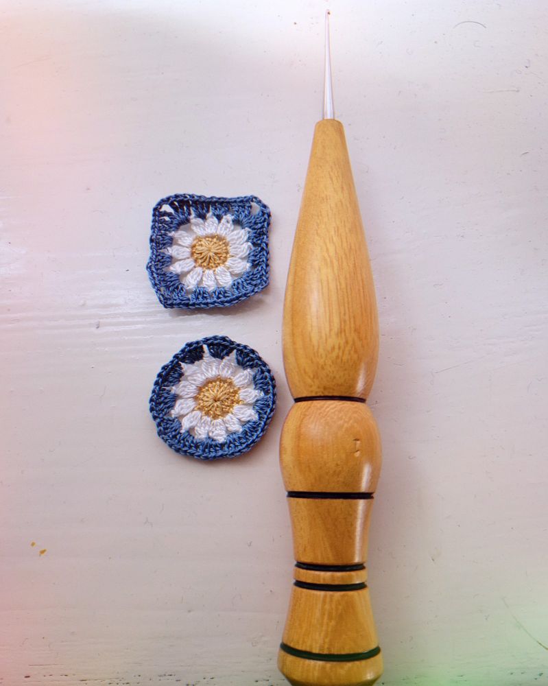 2 micro crochet flower, one square and one circle in blue, white and gold, next to a 1mm crochet hook with a woodturned handle 