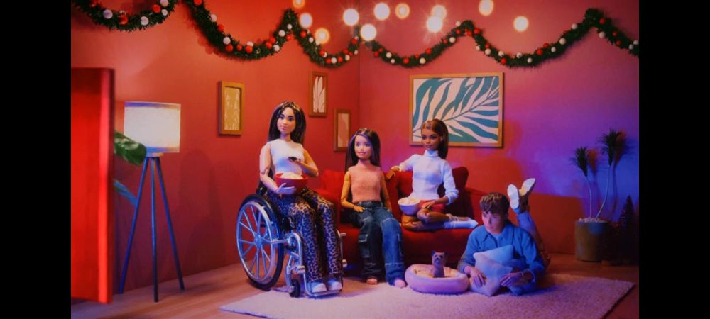 Four Barbie dolls sitting down in the living room watching TV. The one on the left is in a wheelchair.