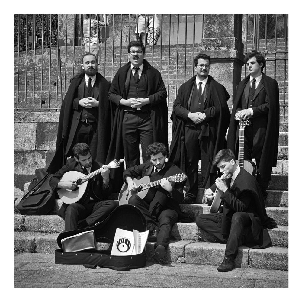 The picture shows seven musicians standing or sitting on a staircase. One of them is singing and three are playing string instruments. All of them are wearing black cloaks. The square photo is black and white and has a white frame.