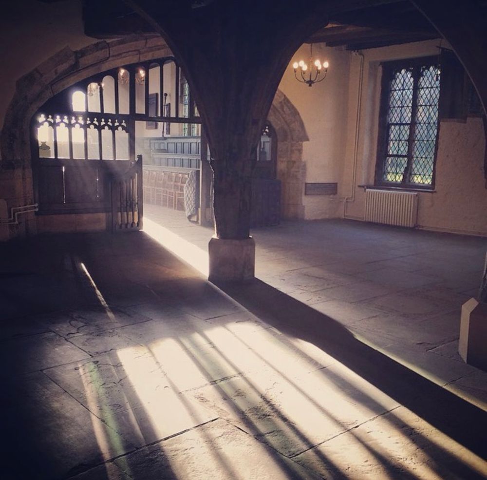 We are looking at a large room with a chapel in the background, it is divided by an oak screen with pointed arches. Sun streams through an unseen window creating misty hazy light and long shadows on the stone flagged floor of the room. 