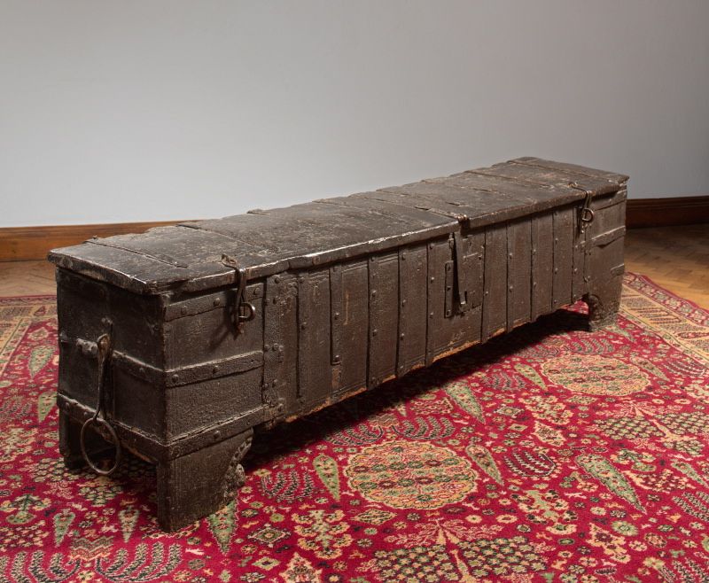 We are looking at a large oak chest which is bounded in iron and has two iron hasps on either end of the lid and then a larger hasp in the centre with a lock. It has a round iron chain handle. 