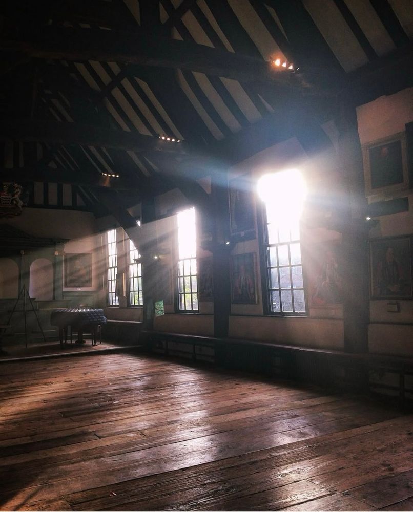 We are looking at a large room with a timber framed roof. There are four Georgian sash windows with light brightly shining though them casting rays of sunshine into the dark room. 