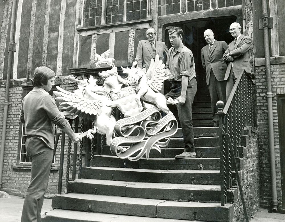 We are looking at a black and white photo of two men carrying a large carved oak Coat of Arms up the stairs into a timber framed building. Three men in suits watch on from the top of the stairs. 