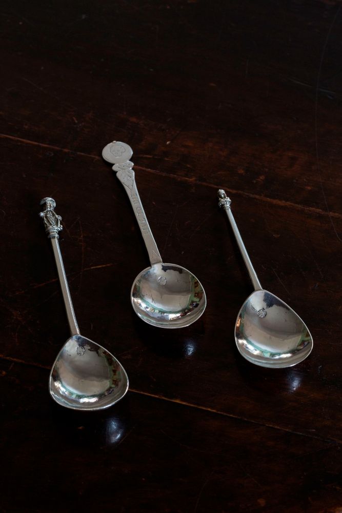 We are looking at three silver spoons on a dark oak table. Two have small figures on the top of them whilst the handle is engraved with a small skull. 