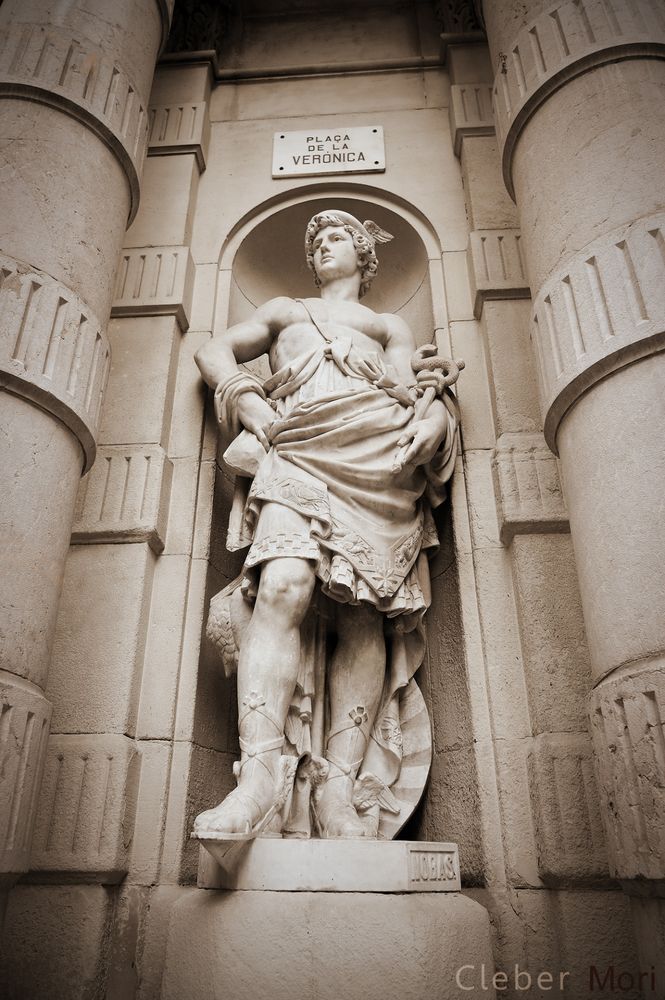 A photo of a statue of the god Hermes, in an alcove between two pillars. He wears a loose wrap around his body. One hand is braced on his hip, and the other bears his caduceus. He wears his winged hat and wrapped sandals.
