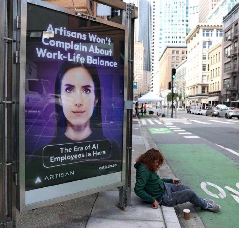 The image shows a juxtaposition of technological advancement and socioeconomic disparity. On the left, an advertisement promotes AI employees, with the slogan: “Artisans Won’t Complain About Work-Life Balance. The Era of AI Employees Is Here.” The ad features a futuristic-looking, digitally rendered woman’s face and the logo of the company “Artisan.” On the right, a disheveled individual appears to be homeless, sitting on the sidewalk next to a green bike lane, holding a small item in their hand, with a disposable coffee cup placed on the ground. In the background, tall urban buildings and city traffic emphasize the urban setting. The image starkly contrasts the promise of AI-driven innovation with the reality of human hardship.