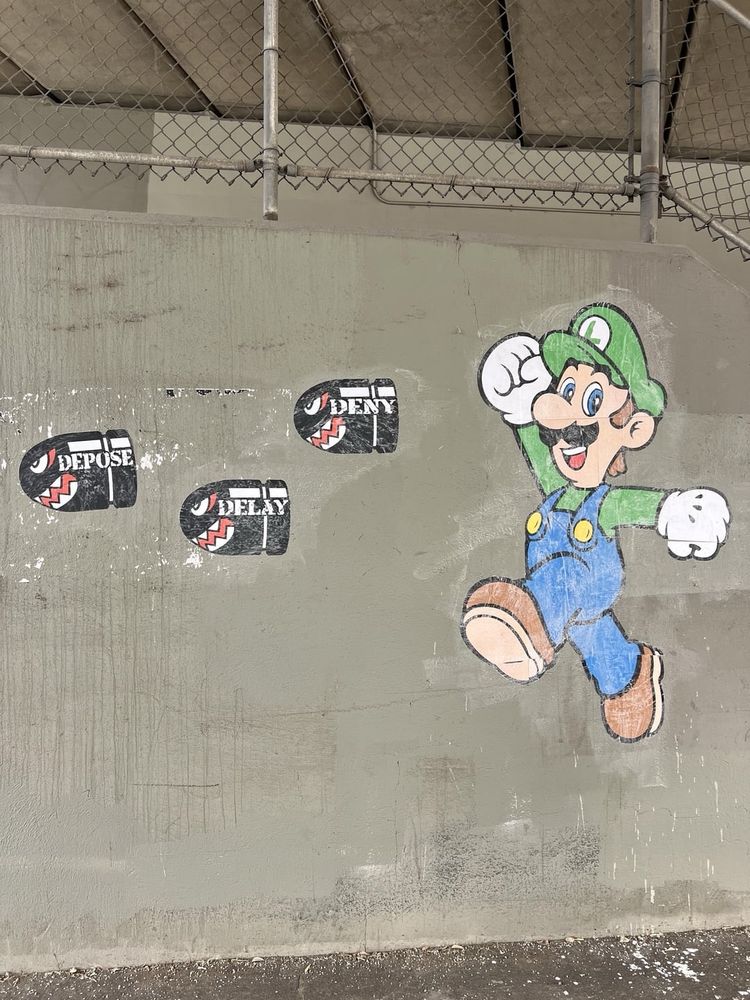 A tall concrete wall beneath an overpass covered by a chain-link fence, featuring street art. On the right side, a hand-drawn cartoon character resembling Luigi from the Mario series leaps upward with one fist raised, wearing his signature green cap, green shirt, blue overalls, and brown boots. To the left, a series of black, bullet-shaped stencils are painted on the wall, each bearing white text such as “DEPOSE”, “DELAY”, and “DENY,” arranged in a line.