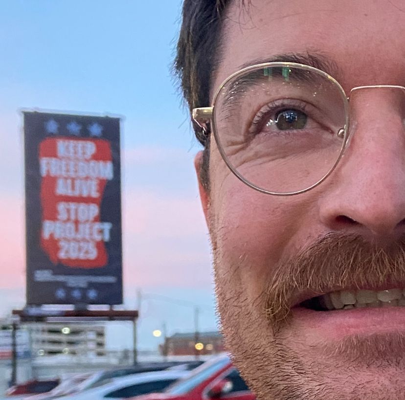A very close crop of Hrothmar’s face on the right. A parking lot of cars is behind them with a large billboard that reads “keep freedom alive, stop project 2025”