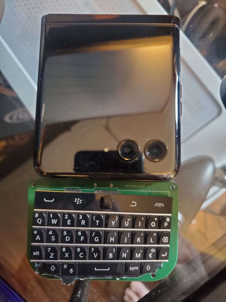 Picture of a folded Motorola Razr 40 Ultra and a BlackBerry keyboard ontop of a pcb sitting next to each other ontop of a glass table.