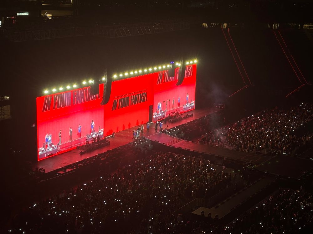 picture a stage with ateez from the ‘in your fantasy’ 2025 tour. the screen onstage is black with the member in the foreground. a very large crowd is in front of stage. 
