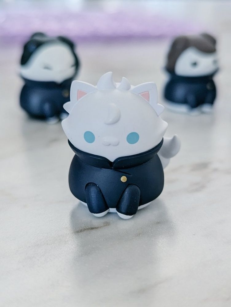 Gojo Satoru cat figurine, commonly referred to as "catoru." Behind him is Suguru Geto (sugucat) and Shoko Ieiri (shocat) out of focus, looking to the right.