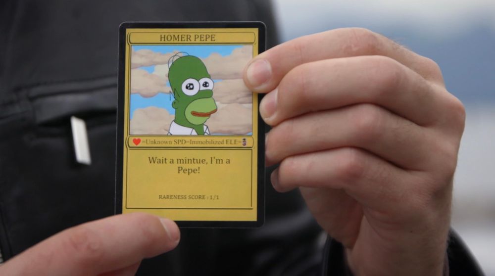 Another screenshot from the film ‘Feels Good Man’ which shows the man from the previous photo holding a really expensive and rare “Homer Pepe” card that he’s spent a daft amount of money buying. It looks like a Pokémon card but with Homer Simpson looking like Pepe the Frog, all green with red lips. 
