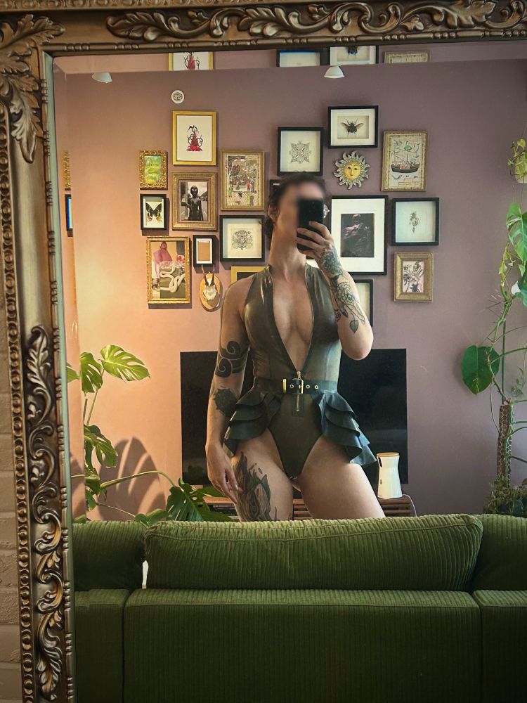 Mistress Kay stands in front of a mirror wearing a translucent high cut latex bodysuit and black latex peplum skirt. 