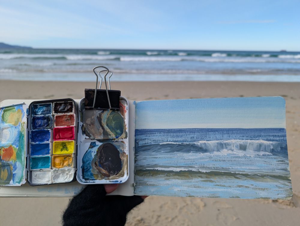 I'm holding my sketchbook up with the beach in the background. Clipped to my sketchbook on the left page is my plein air gouache palette  with 12 colours. On the right page is a gouache study of the breaking waves.