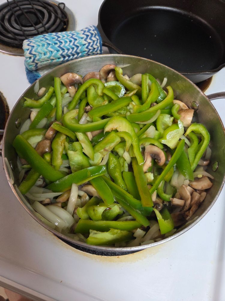 A deep saute pan filled with onions peppers and mushrooms.