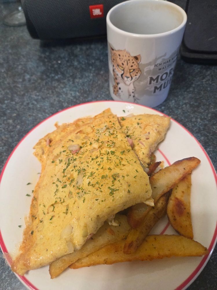 A plate with a folded omelette, cut in half and garnished with a little parsley. Next to the omelette are some potato wedges. Next to the plate is my coffee cup with a cartoon grumpy cheetah.