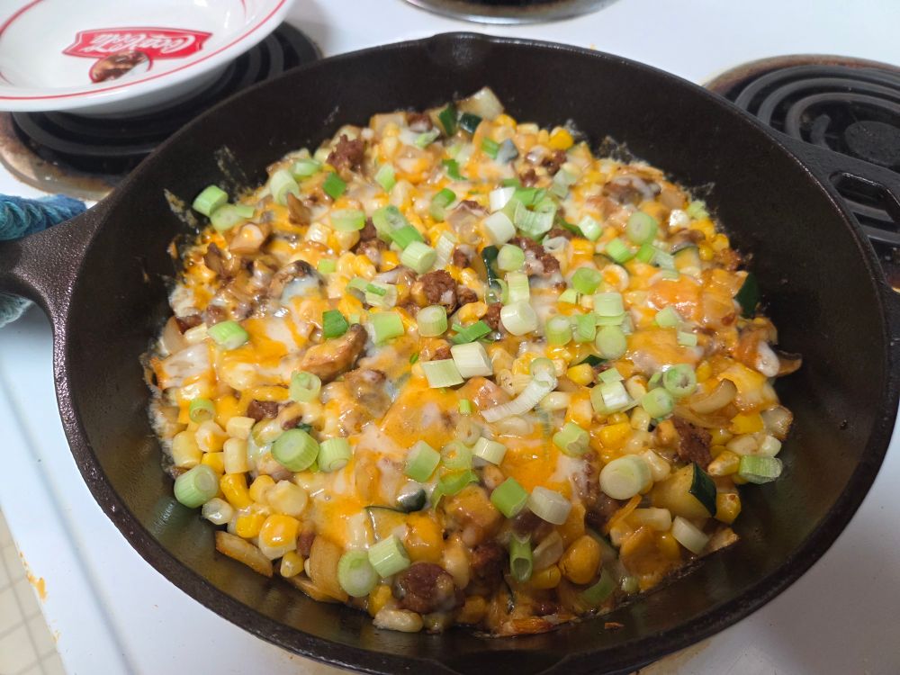 A cast iron skillet filled with a mix of chorizo, zucchini, onions, mushrooms and corn. It's topped with melted cheese and green onions. 