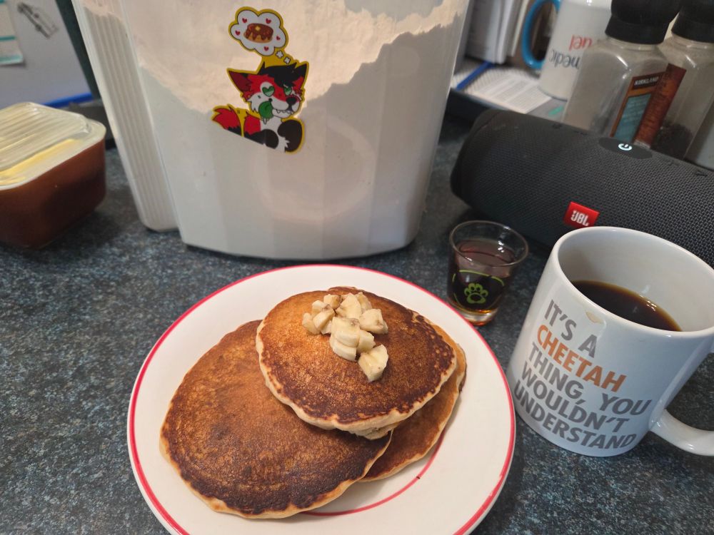 A short stack of pancakes topped with some chopped up bananas. Next to it is a shot glass from VF filled with maple syrup, a coffee mug that has "it's a cheetah thing; you wouldn't understand" on it, and my bin of pancake mix with a sticker of a pancake yote on it. 