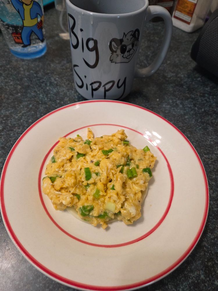 A plate with some soft scrambled eggs with green onions mixed in. Next to it is a blue mug that says "Big Sippy" and a silly sketch of my cheetah on it. 