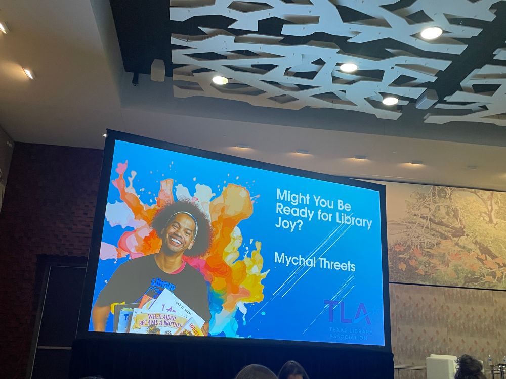 Photograph of the title slide for a slide deck showing Mychal Threets standing behind a display of picture books. The title text reads: Might You Be Ready For Library Joy? The TLA logo is placed on the bottom right corner. 