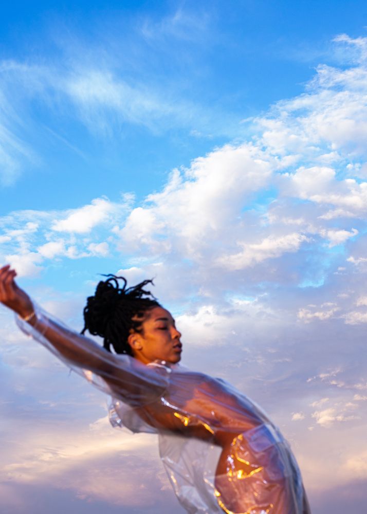 Color Photo of woman in a transparent plastic outfit posing with a blue, cloudy sky backdrop. The woman is slightly out of focus. 