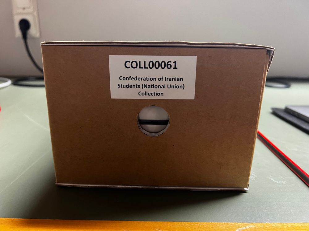 A small brown archival storage box with a white label reading “COLL00061 – Confederation of Iranian Students (National Union) Collection.” The box rests on a green desk near a wall outlet in a workspace for archival processing.