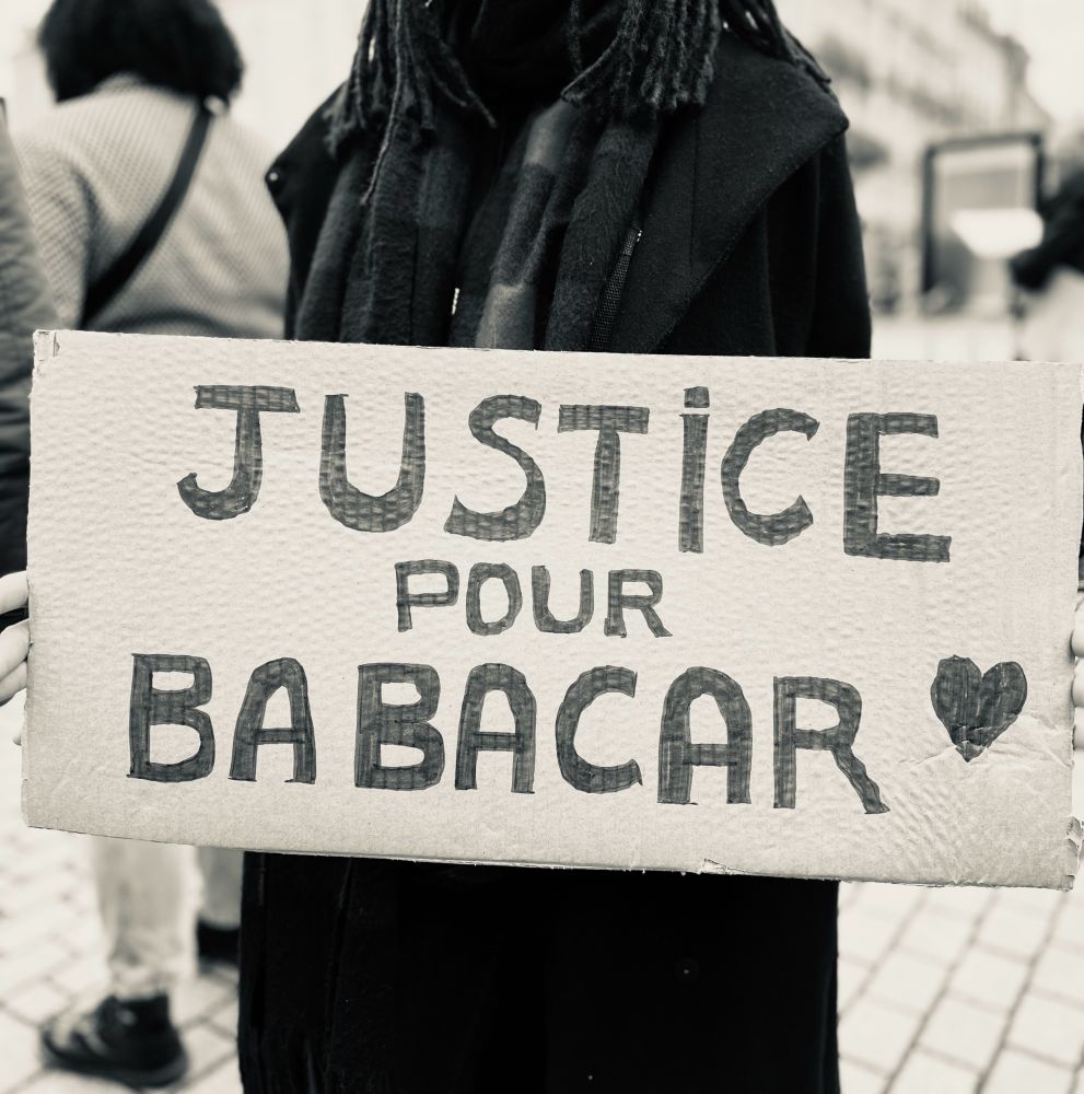 2. Troisième image : Un gros plan d’une pancarte en carton tenue par une personne vêtue de noir. Le texte écrit à la main indique : “Justice pour Babacar,” accompagné d’un petit cœur dessiné dans le coin inférieur droit.