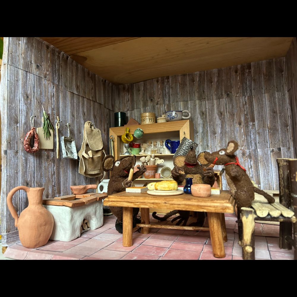 Photo of Minimus, Minima and Silvius, the Latin mice, in their newly redone kitchen. There's a big plate on the new wooden table with a whole cheese, and Minimus is holding a cheese knife. There are pegs above the stove with a chopping-board, a muffin pan, a sprig of rosemary and a salami style sausage. 